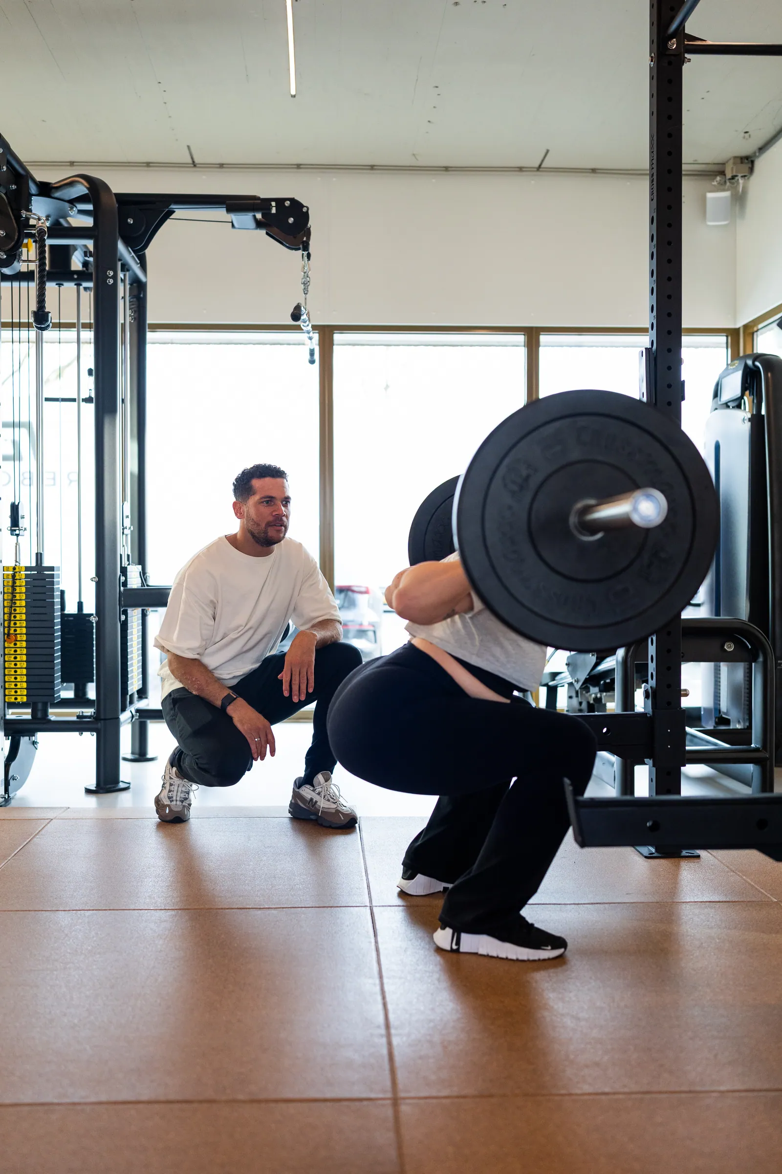 Coach knielt naast atlete die squat doet met barbell bij ramen.