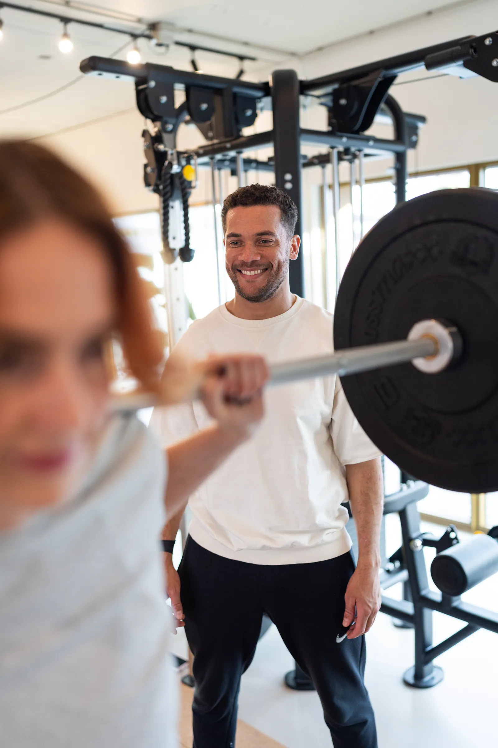 Coach lacht terwijl atlete op voorgrond met barbell uit beeld is.