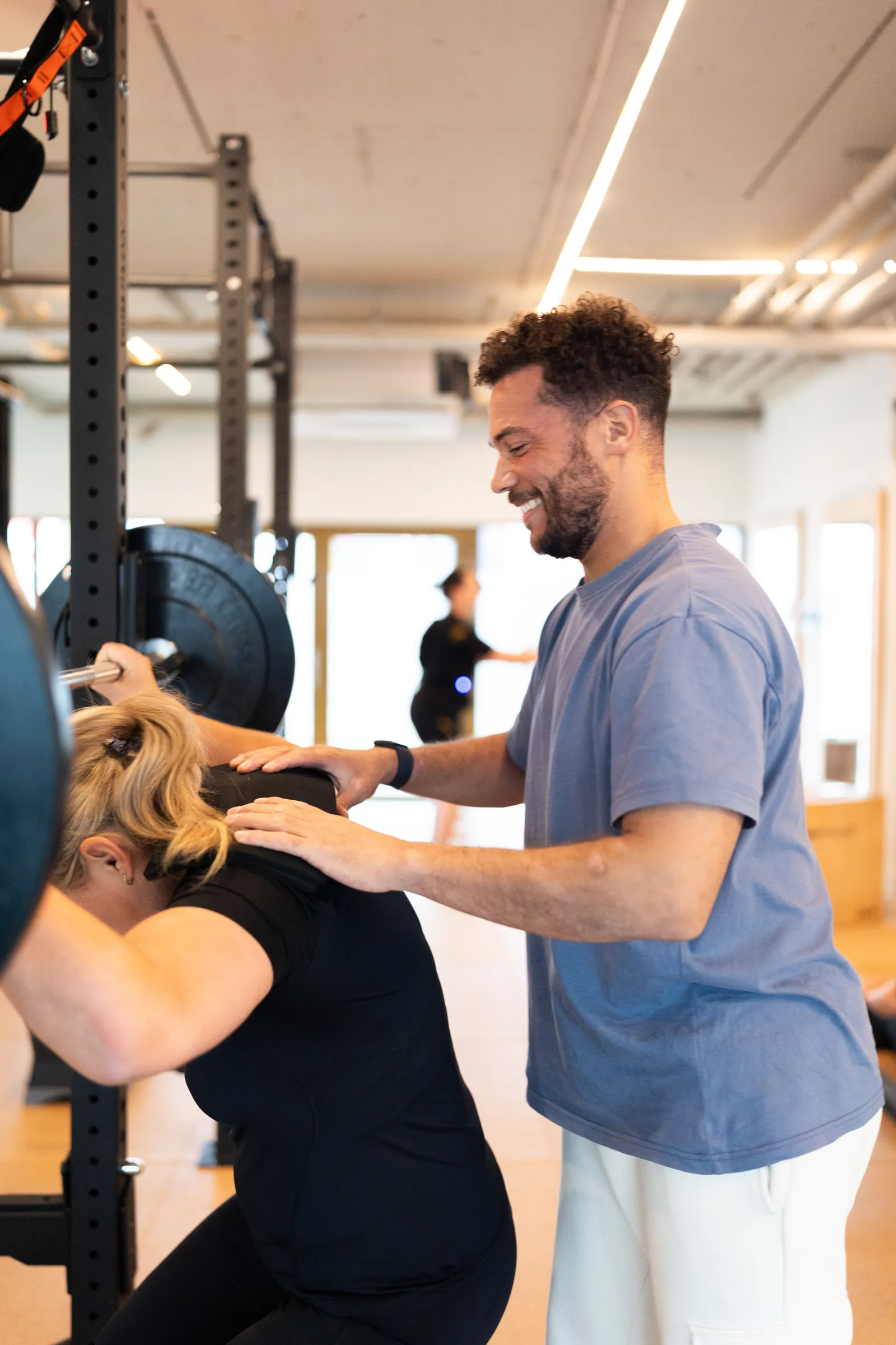 Lachende coach spot vrouw tijdens squat; handen op schouders, rek links.